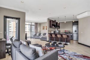 Spacious open-plan living room and kitchen with breakfast bar, lounge area, workspace, balcony access and modern amenities in Cape Town CBD apartment.